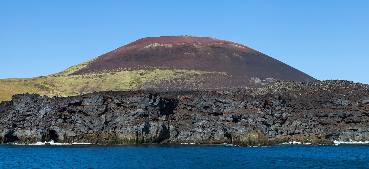 Eldfell’s red and dark volcanic cone rises above a black lava shoreline and blue sea.