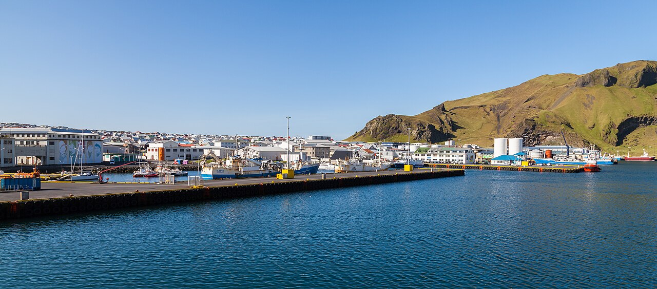 Fishing boats lie beside the waterfront of Heimaey, with buildings and a green cliff behind the harbour.