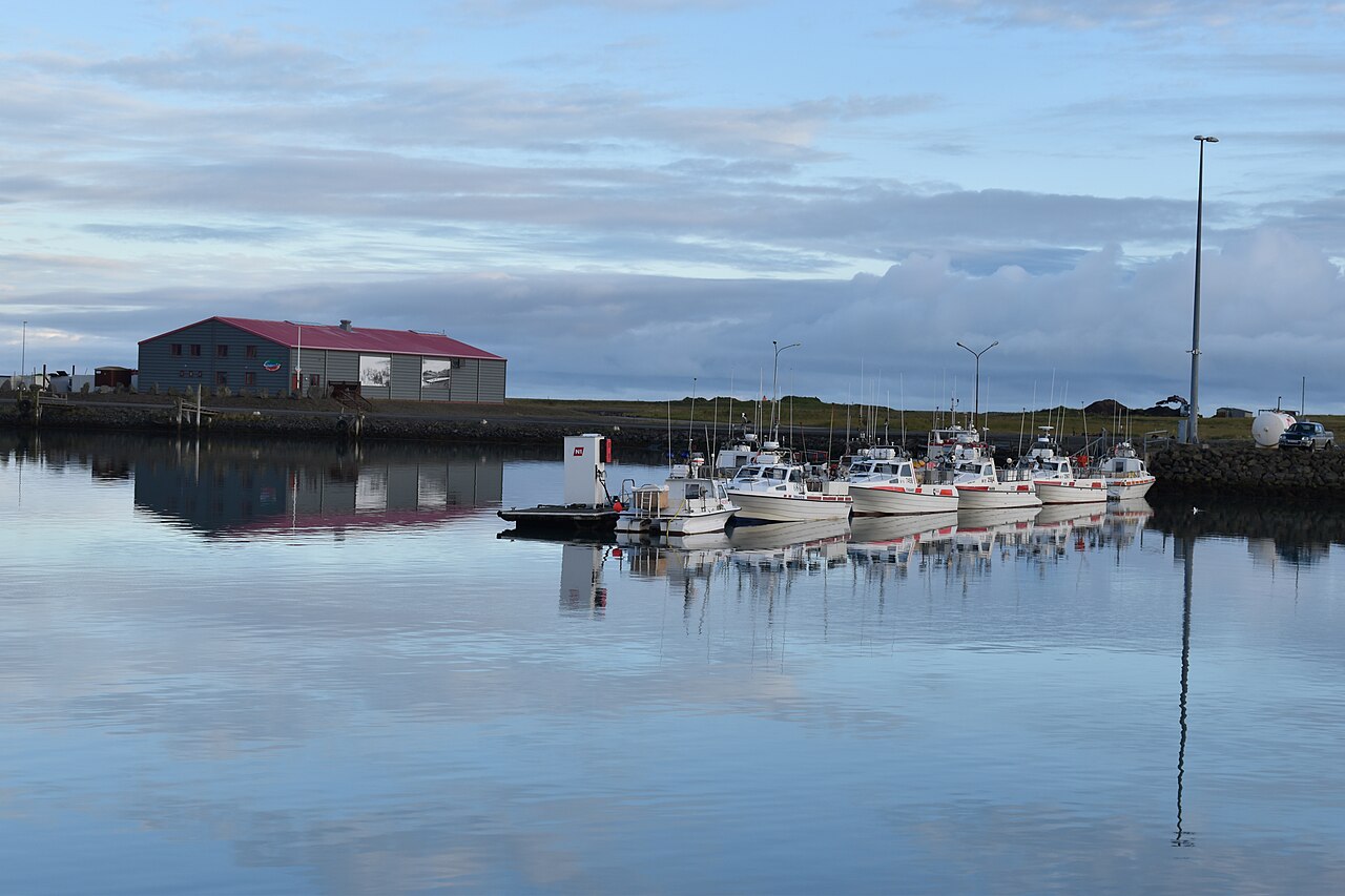 A row of small fishing boats and a red-roofed waterfront building reflect in Höfn’s calm harbour.