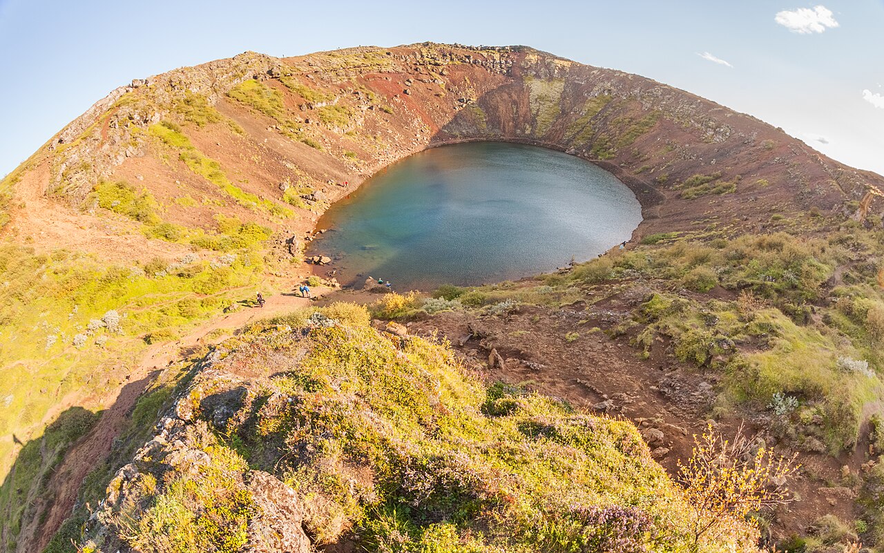 A blue-green lake fills Kerið’s bowl-shaped crater, surrounded by reddish rock and low vegetation.