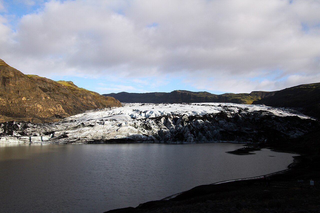 The crevassed ice front of Sólheimajökull meets a dark lake between rocky valley walls.