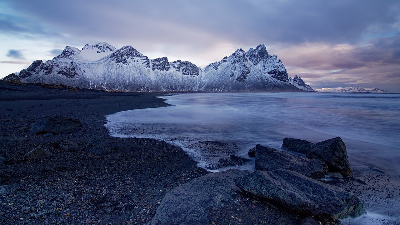 Snow-covered Vestrahorn rises behind a dark beach and shallow coastal water under a cloudy winter sky.