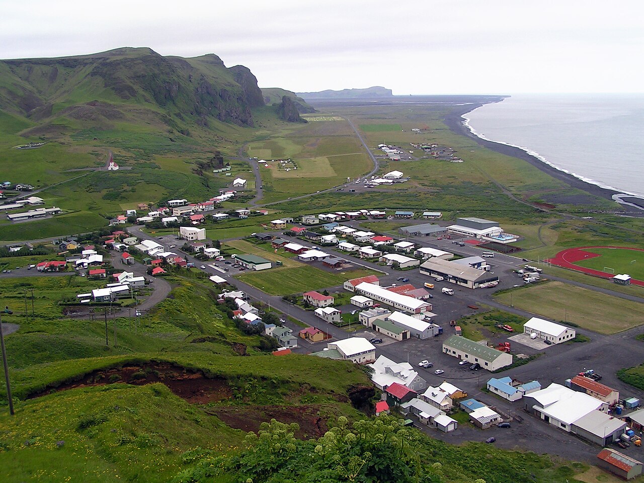Vík’s houses and roads spread below green cliffs beside a long black-sand coastline.